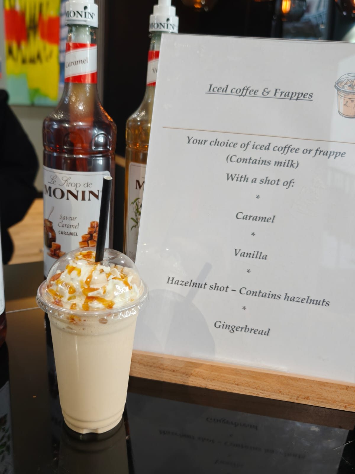 Caramel frappe topped with whipped cream and caramel drizzle next to the syrup menu