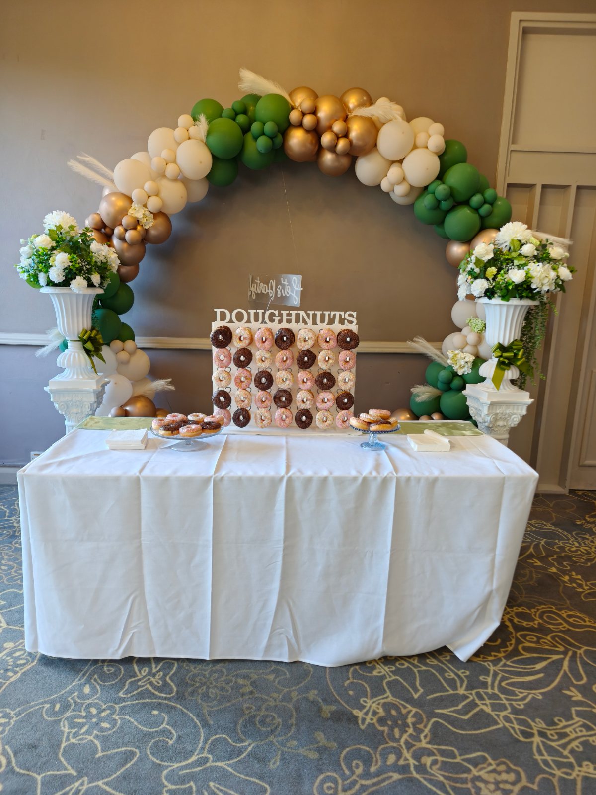 Doughnut wall filled with iced ring doughnuts on a dressed table under a balloon arch