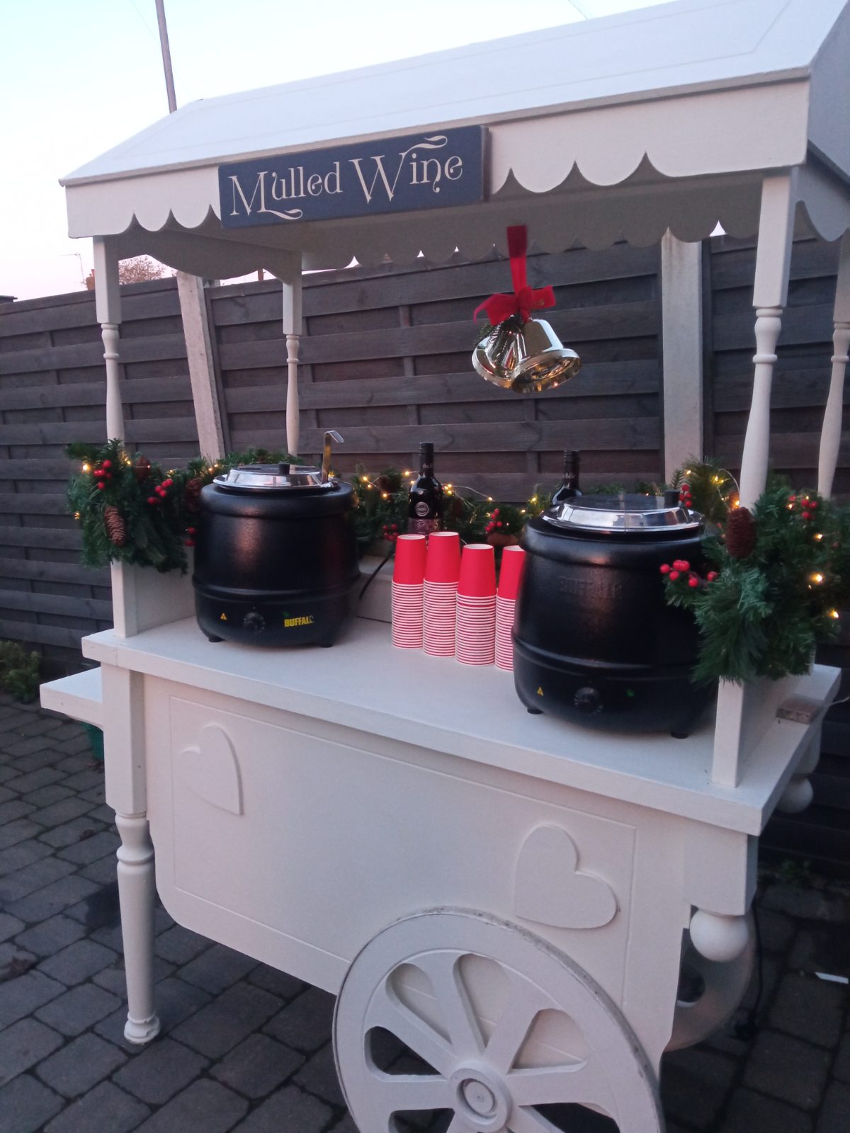 White mulled wine cart with two urns, red cups and Christmas garlands